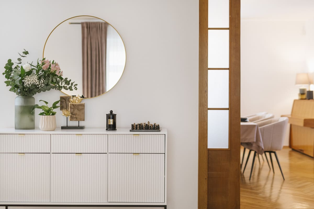 Biała komoda, nad nią okrągłę lustro, z prawej pokój z pianinem oraz kremowymi krzesłami